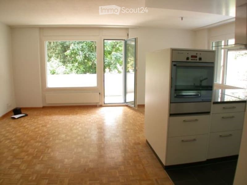 Empty room with wooden flooring, white walls, large windows, and a partially open sliding door leading outside. A small cabinet and oven are visible on one side.
