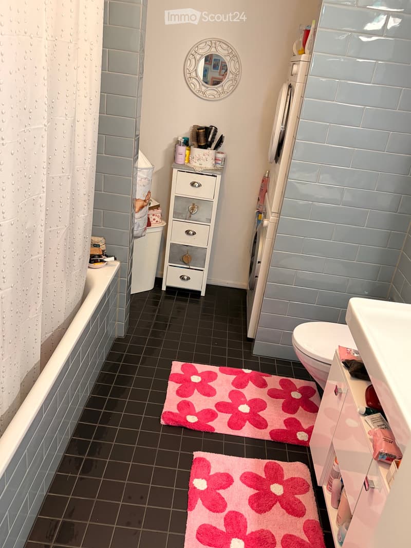 Modern bathroom with light blue tiles, black floor tiles, a bathtub, toilet, mirror, and a small cabinet with toiletries. Pink flower rugs are placed on the floor.