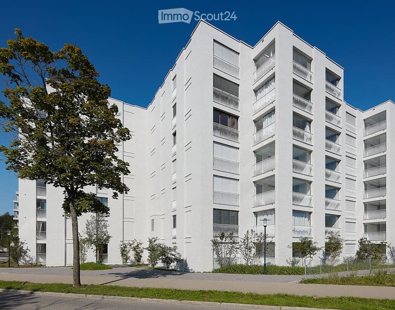 Modern white apartment building with multiple balconies and large windows, surrounded by greenery and a paved area.