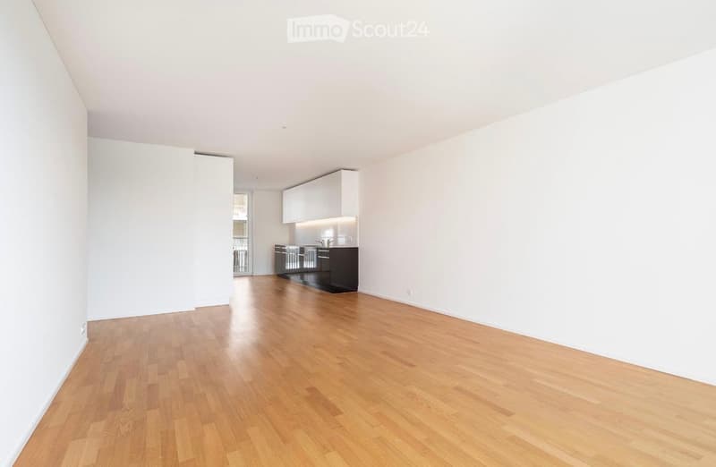 Empty room with wooden flooring, white walls, and a small kitchen area with cabinets and appliances visible in the background.