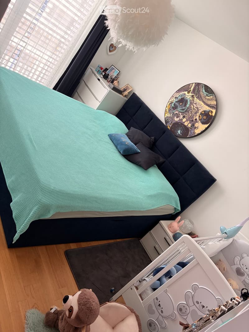 A cozy bedroom with a bed covered in a light green blanket, pillows, a nightstand with various items, a mirror, a plush toy, and a small chair with a cushion.