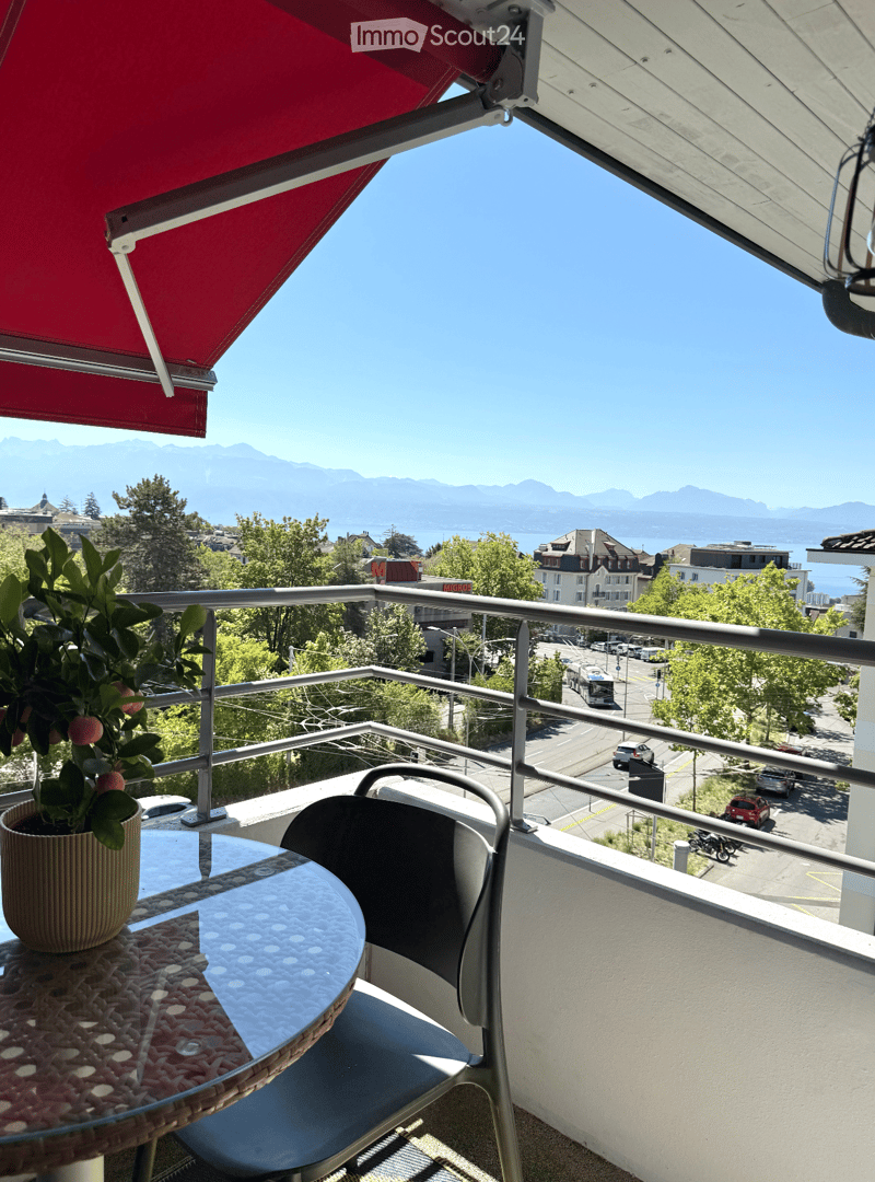 Balcony with a view of a cityscape and mountains, featuring a table, chair, and potted plant.