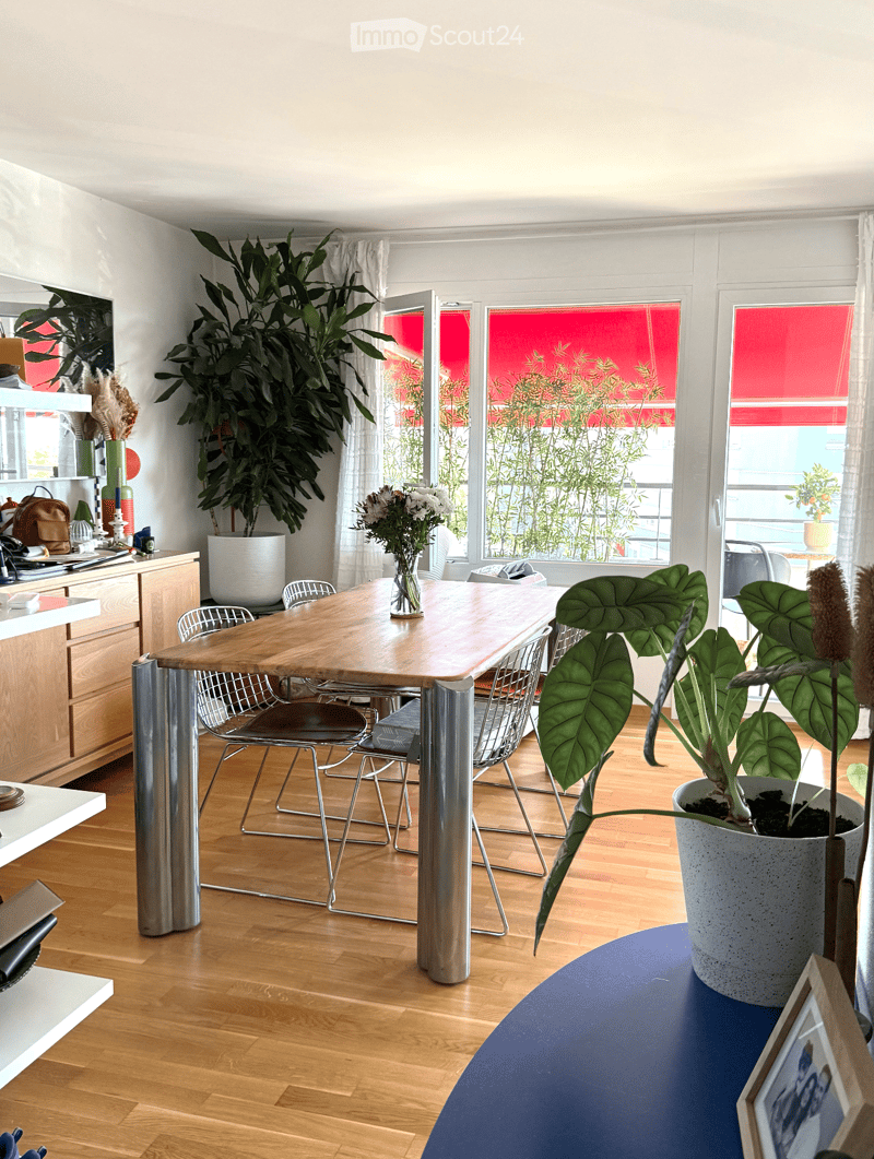 Bright, modern dining area with wooden floor, large wooden table, metal chairs, potted plants, and a vase with flowers on the table.