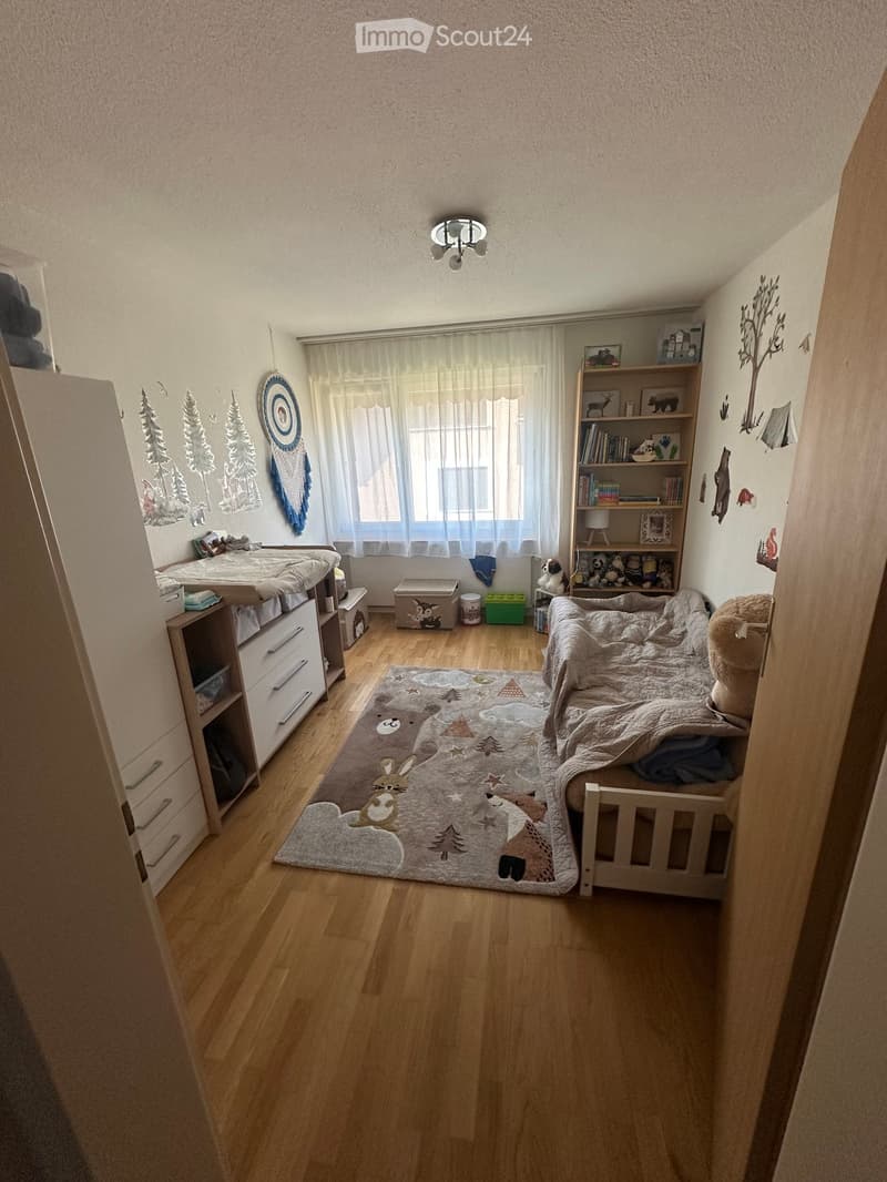 Cozy children's bedroom with wooden flooring, a bed with a blanket, a bookshelf, a drawer unit, a decorative rug, and wall decorations.