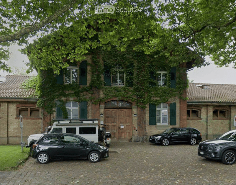 A two-story brick building with green shutters and a large wooden door, surrounded by trees. Several cars are parked in front of the building.