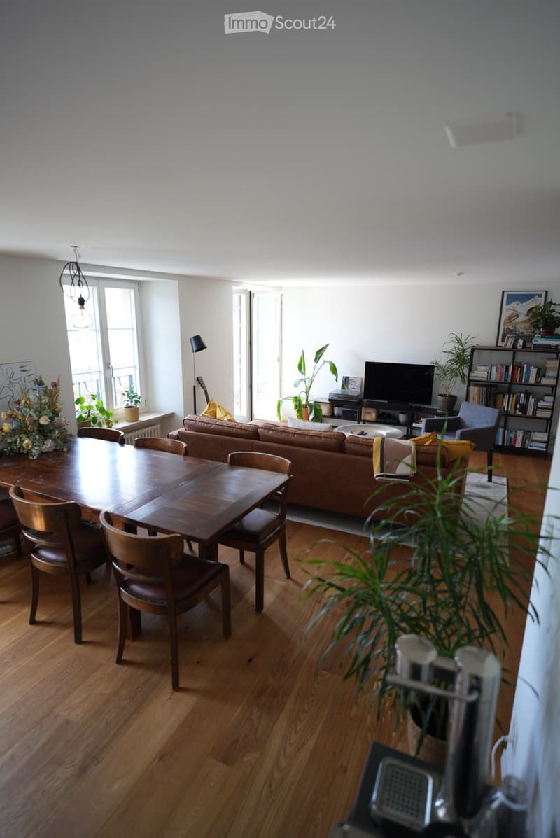Bright living room with wooden flooring, a brown leather sofa, a wooden dining table with chairs, a TV, bookshelf, and decorative plants.