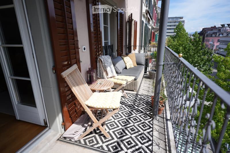 Balcony with a grey sofa, wooden folding chair, small table, patterned rug, and potted plants, overlooking a cityscape.