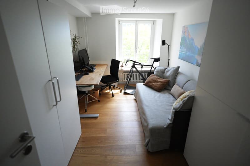 A compact, well-lit room with a sofa, desk, and bicycle. The space features a wooden floor, large windows, and a mix of modern and cozy furnishings.