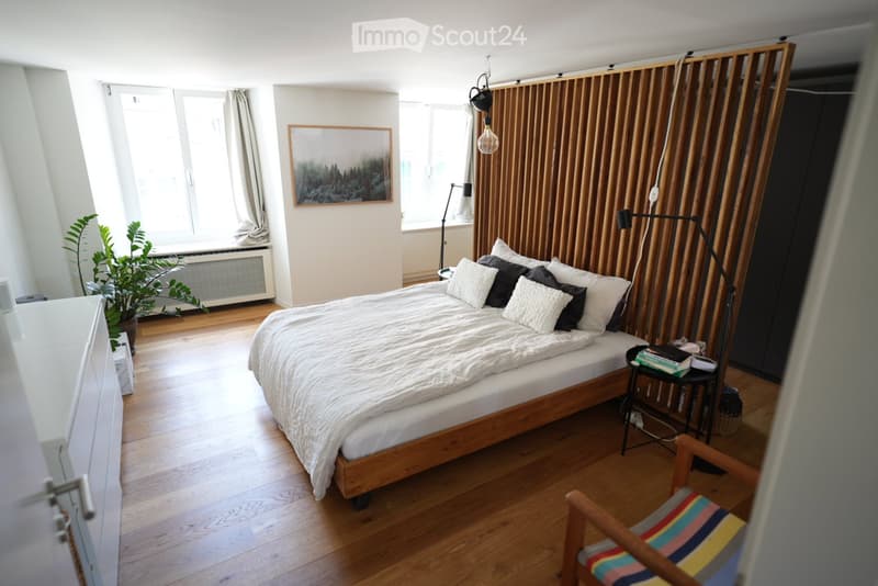 Cozy bedroom with wooden flooring, a bed with multiple pillows and a duvet, a nightstand with books, a colorful chair, a potted plant near the window, and a picture frame on the wall.