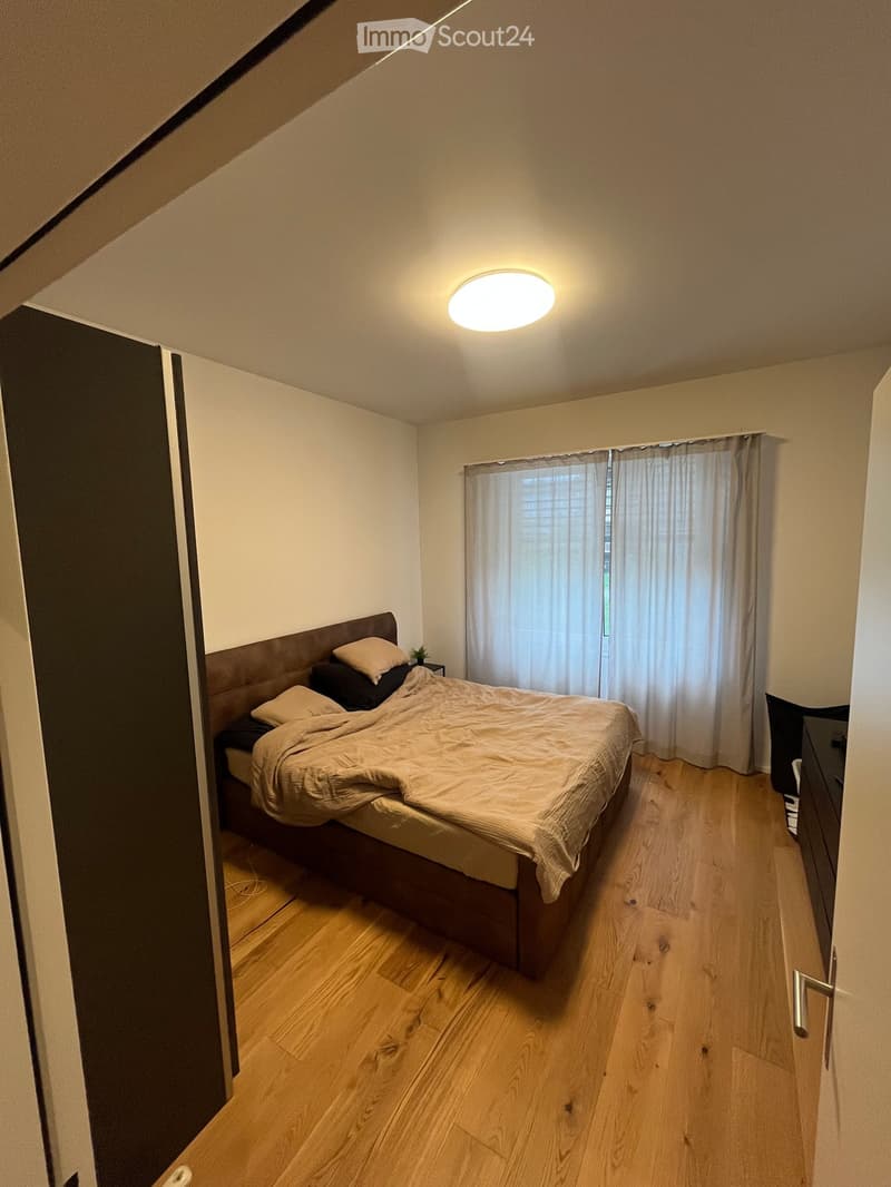 Cozy bedroom with wooden flooring, a large bed with pillows and a blanket, a nightstand with a small plant, and a window with white curtains.