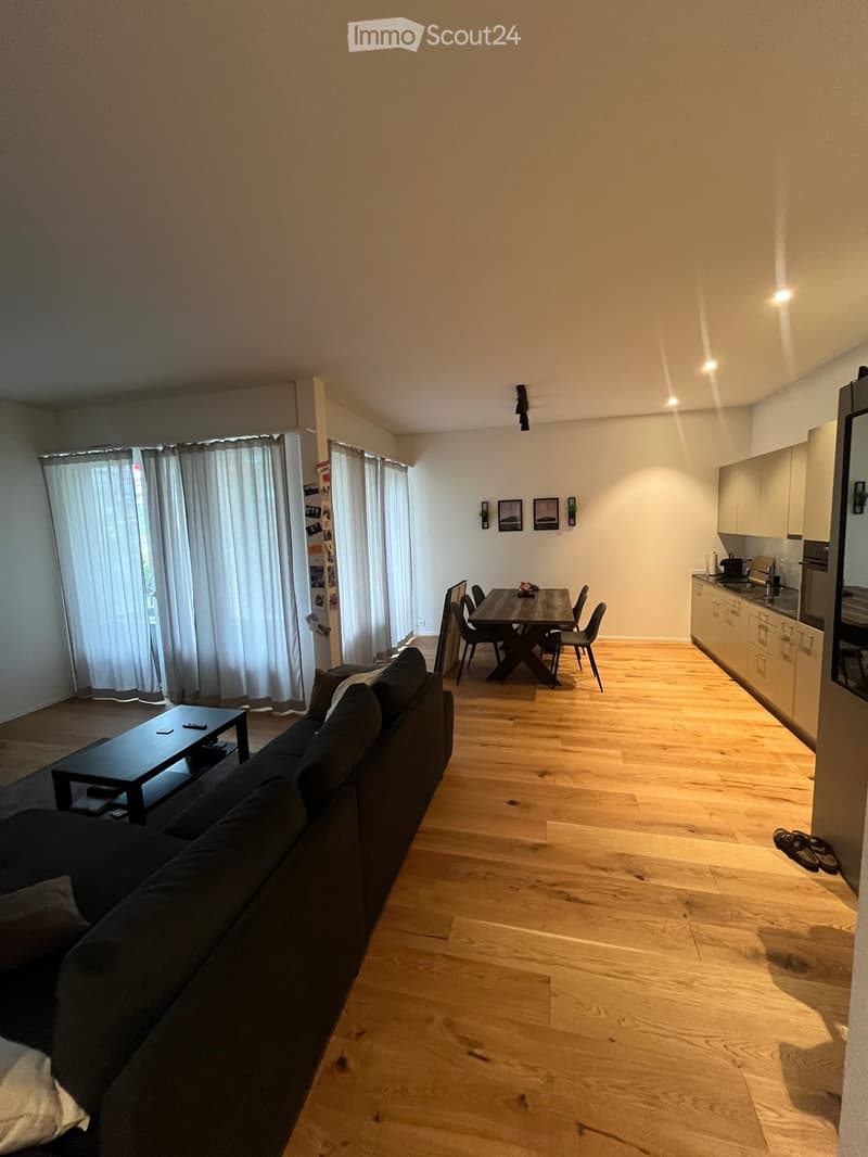 Open-plan living and dining area with wooden flooring, a dark grey sofa, coffee table, dining table with chairs, and a modern kitchen with cabinets and appliances.