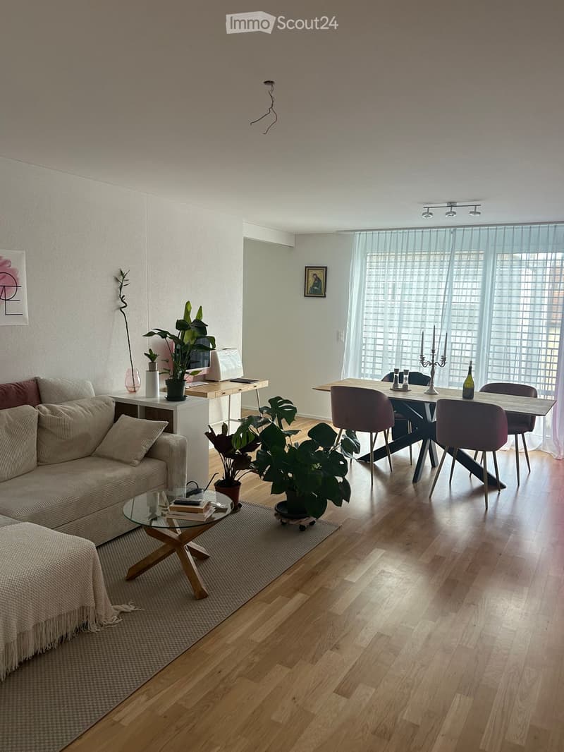 A modern living room with a sofa, coffee table, dining area, and several plants. The room features wooden flooring, large windows with sheer curtains, and a mix of contemporary and minimalist decor.