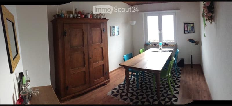 A furnished room with a wooden table, colorful chairs, a large wooden cupboard, and various decorative items on the walls and surfaces.