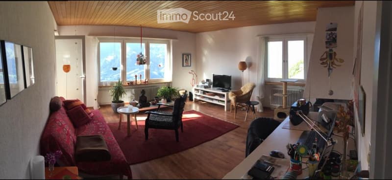 Bright living room with wooden floors, large windows offering a scenic view, a red sofa, a coffee table, a wicker chair, a cat tree, a desk with a laptop, and a table lamp.