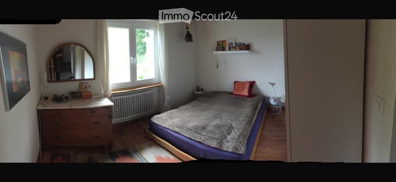 Cozy bedroom with a double bed, dresser, mirror, and shelves. The room features wooden flooring and large windows with white curtains.