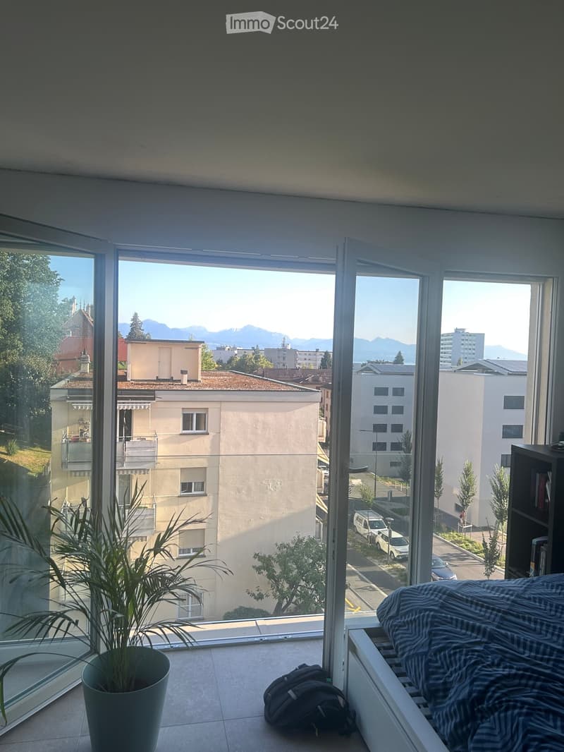 Bright bedroom with large windows offering a scenic view of the city and mountains. Features a bed with striped bedding, a potted plant, and a backpack on the floor.