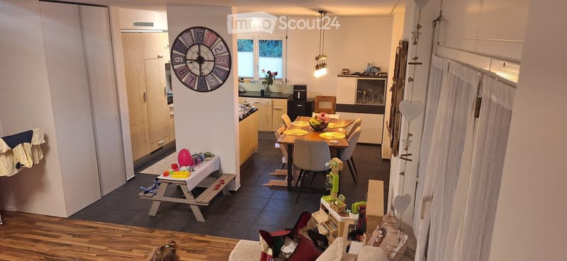 A modern, open-plan living and dining area with a sofa, dining table, chairs, kitchen cabinets, and various decorative items including a large wall clock and a balloon.