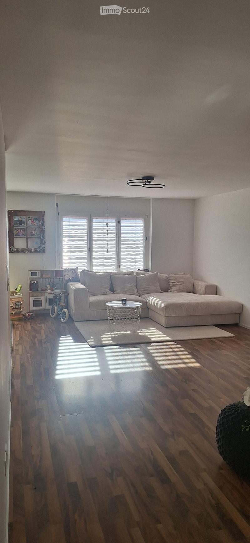Modern living room with a large sectional sofa, coffee table, and wooden flooring. The room features large windows with blinds and a small kitchen area visible in the background.