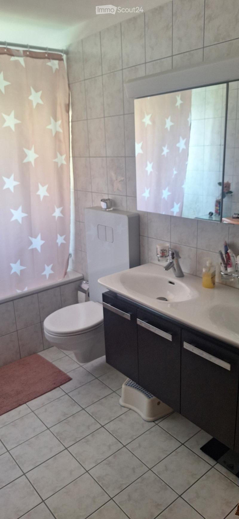 Bathroom with a toilet, sink, mirror, and shower curtain featuring star patterns. The sink has a modern faucet and a small cabinet underneath. A red bathroom rug is placed in front of the toilet.