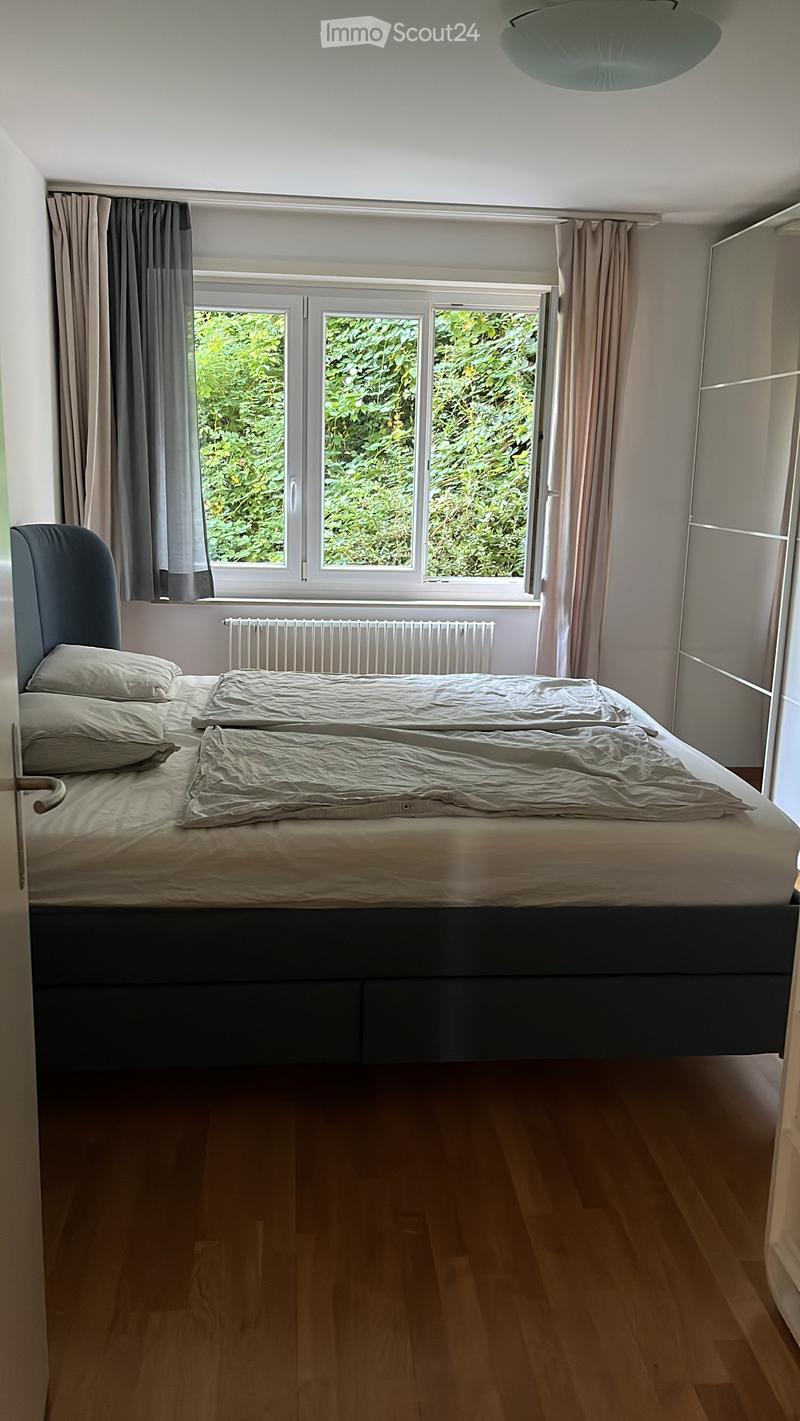 Bedroom with a double bed, pillows, radiator, and large windows with curtains. The room has wooden flooring and a modern aesthetic.