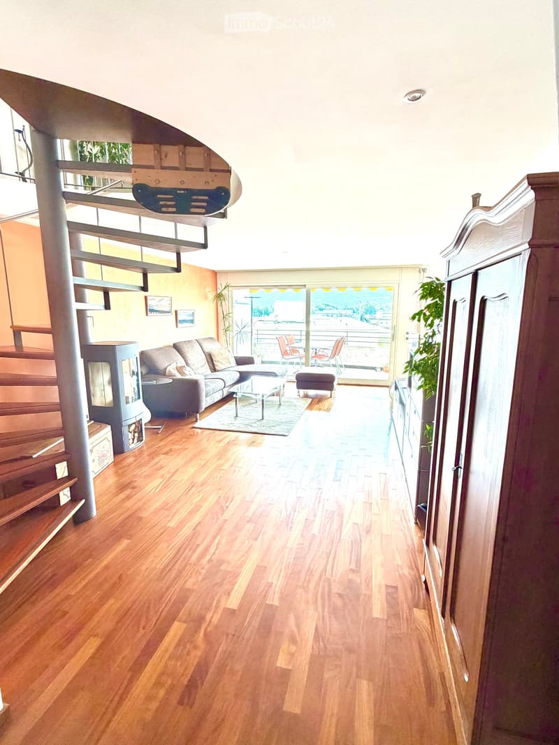 Modern living room with wooden flooring, a grey sectional sofa, coffee table, ottoman, fireplace, and a view of a balcony with outdoor seating.