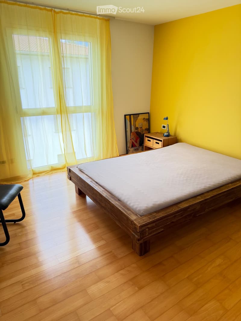 Bright bedroom with wooden flooring, large window with yellow curtains, bed with white mattress, nightstand with lamp and vase, and a black chair.