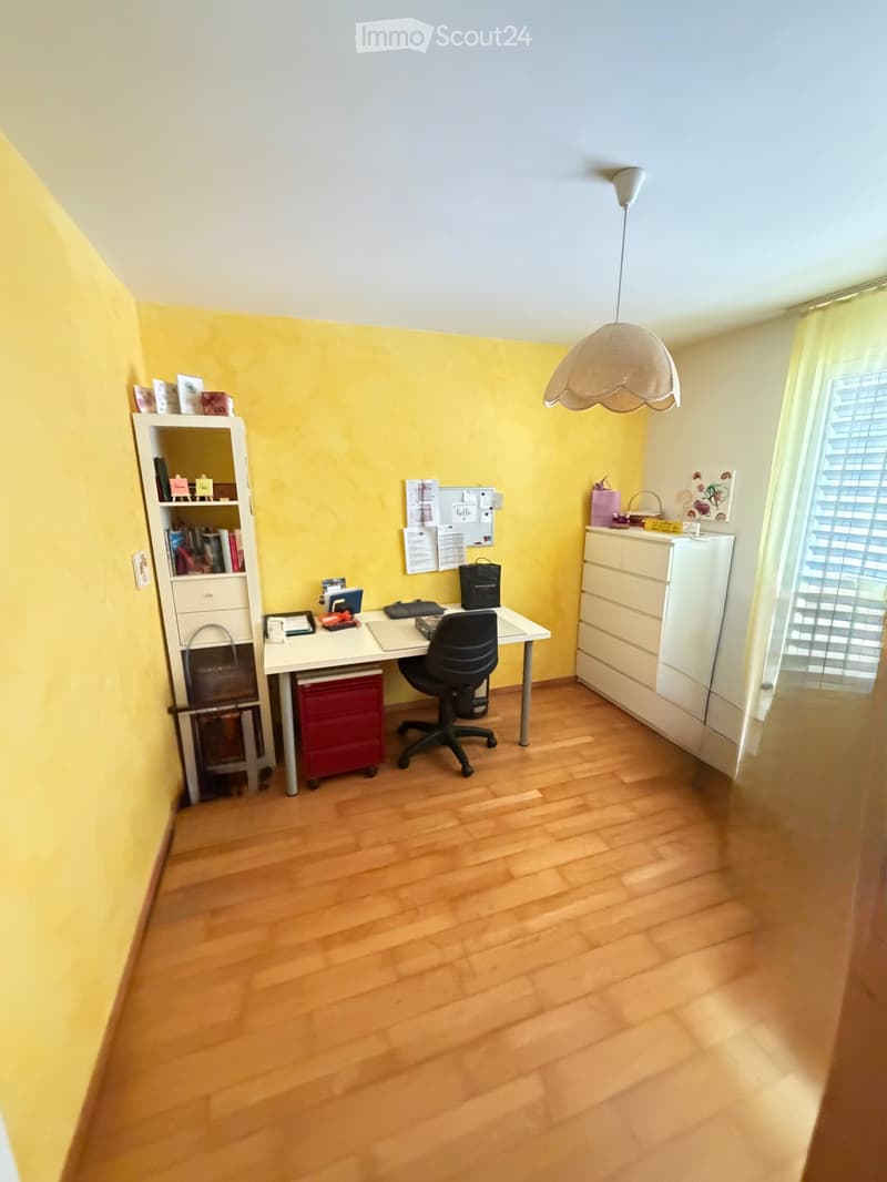 Brightly lit room with wooden flooring, a white desk, black office chair, white bookshelf, and a white drawer unit.