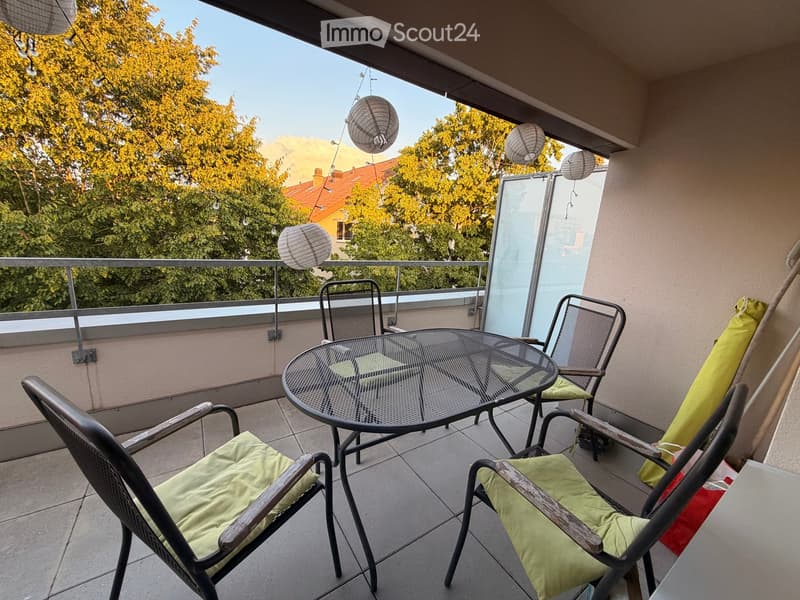 Balcony with a round glass table and four chairs, green cushions, hanging lanterns, and a view of trees and buildings.