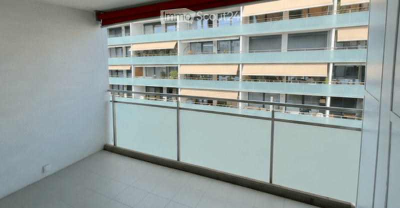 Balcony with modern glass railing, tiled floor, and view of a multi-story residential building with balconies and awnings.