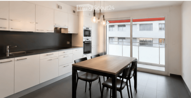 Modern kitchen and dining area with white cabinets, black countertops, a wooden dining table with black chairs, and a large window overlooking a balcony.