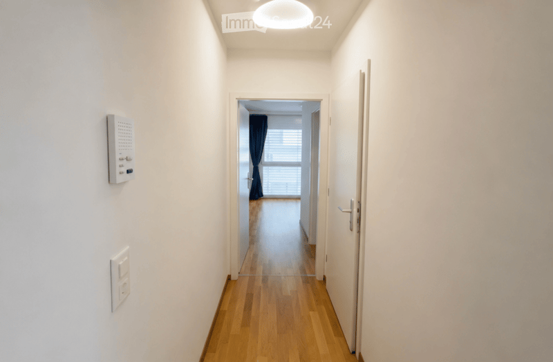 A narrow hallway with light wooden flooring, white walls, and a ceiling light fixture. A door on the right leads to another room with a window and dark curtains.