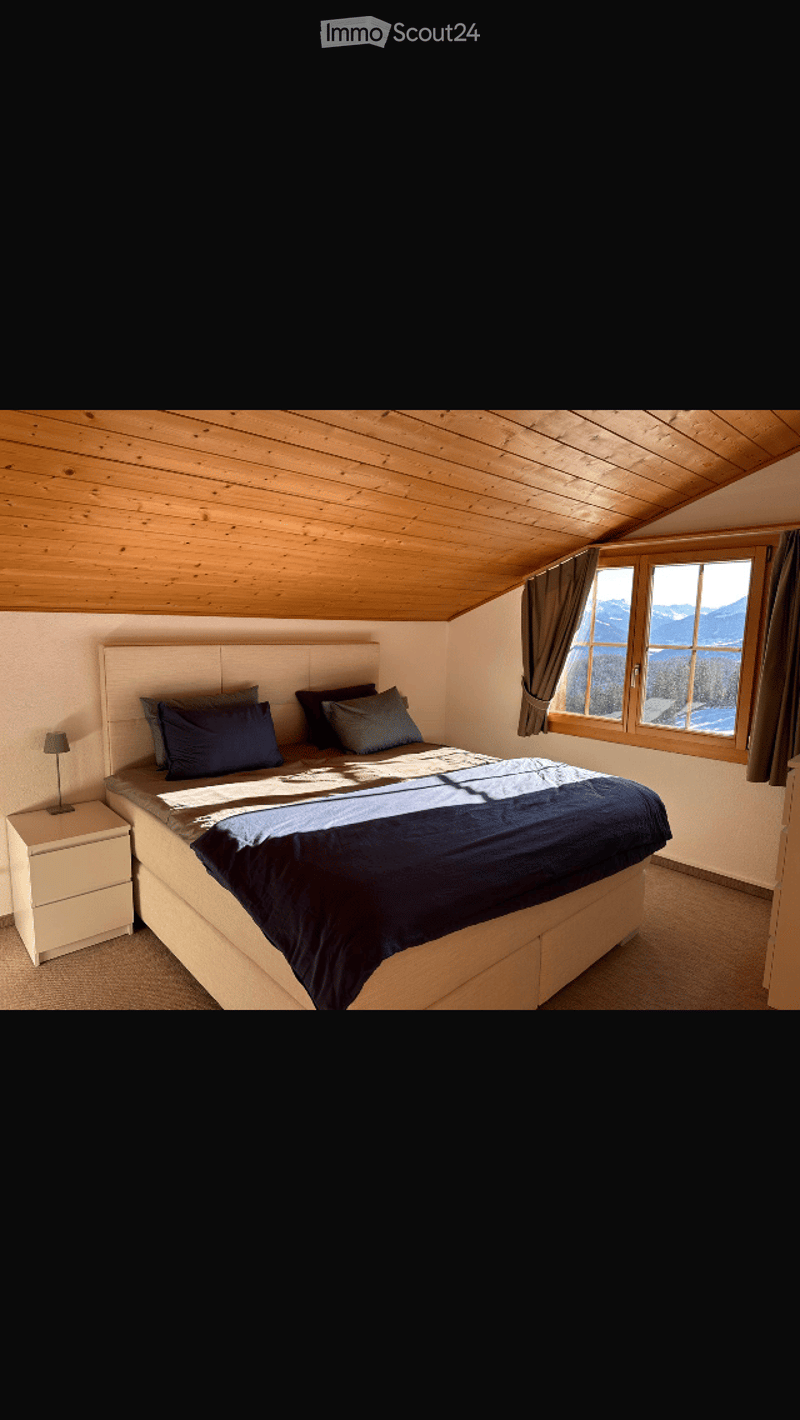 Cozy bedroom with wooden slanted ceiling, large window with mountain view, neatly made bed with pillows and blankets, nightstand with lamp.
