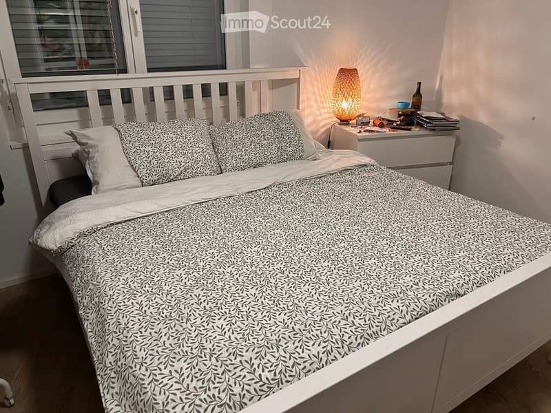 Cozy bedroom with a white bed, patterned bedding, a nightstand with a lamp, and a window with blinds.