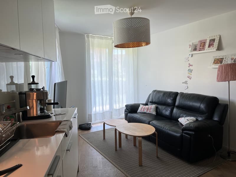 Modern open-plan living and kitchen area with a black leather sofa, wooden coffee table, small wooden stools, a kitchen counter with a coffee machine, and framed pictures on the wall.
