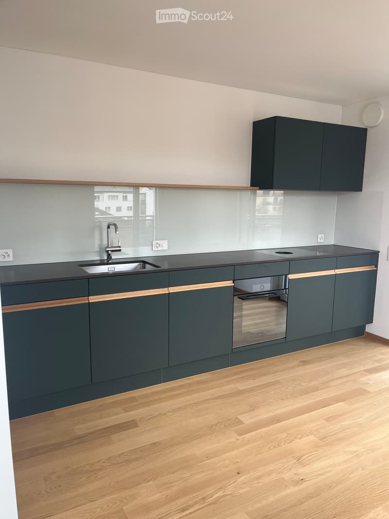 Modern kitchen with dark grey cabinets, wooden accents, a stainless steel sink, and a built-in oven. The backsplash is a sleek, light-colored glass tile.
