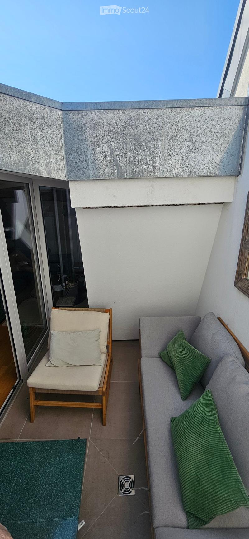 Balcony with a grey sofa, wooden chair, green cushions, and a small green rug.