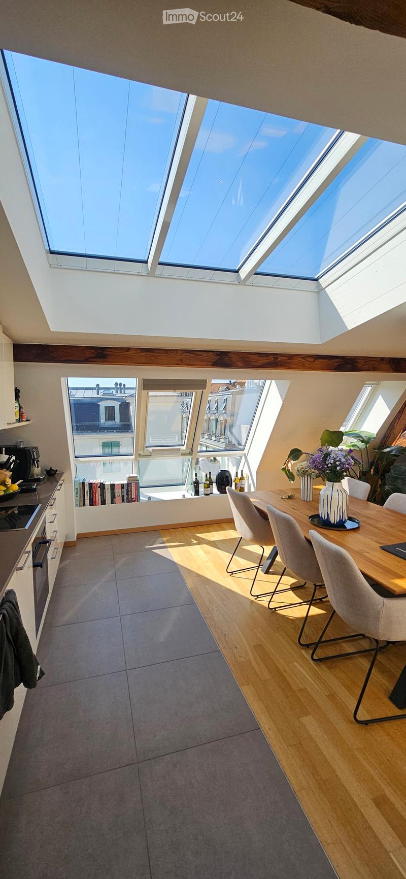 Bright, open-plan room with skylights, wooden dining table, modern chairs, kitchen area with appliances, and decorative vases.
