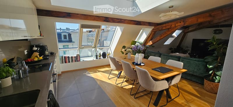 Bright, open-plan living and dining area with wooden floors, large windows, and skylights. Features a green sofa, wooden dining table with chairs, kitchen area with appliances, and decorative plants.