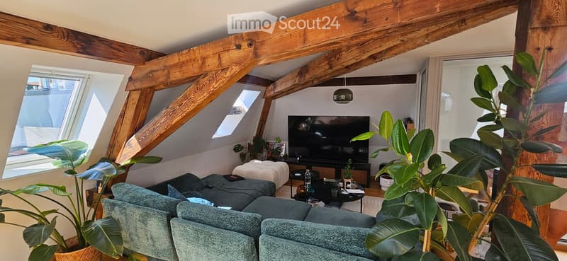 Modern living room with exposed wooden beams, large windows, grey sectional sofa, coffee table, television, and potted plants.