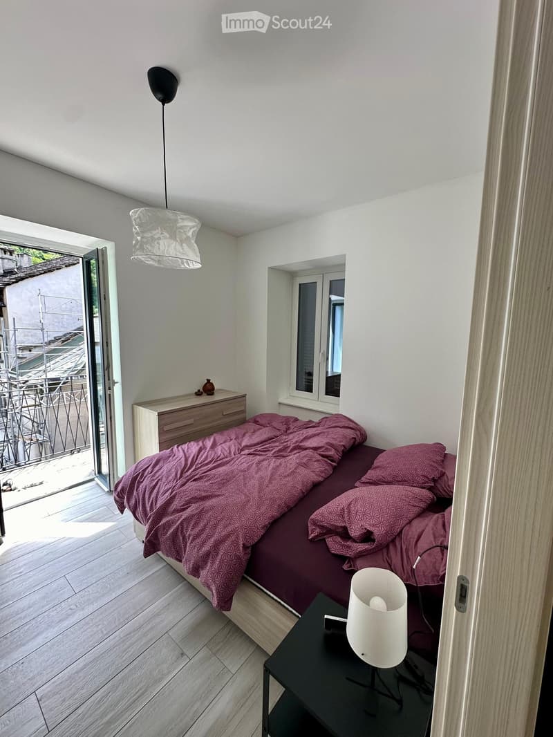 Modern bedroom with a wooden bed, purple bedding, a nightstand with a lamp, a small drawer unit, and a vase on top.
