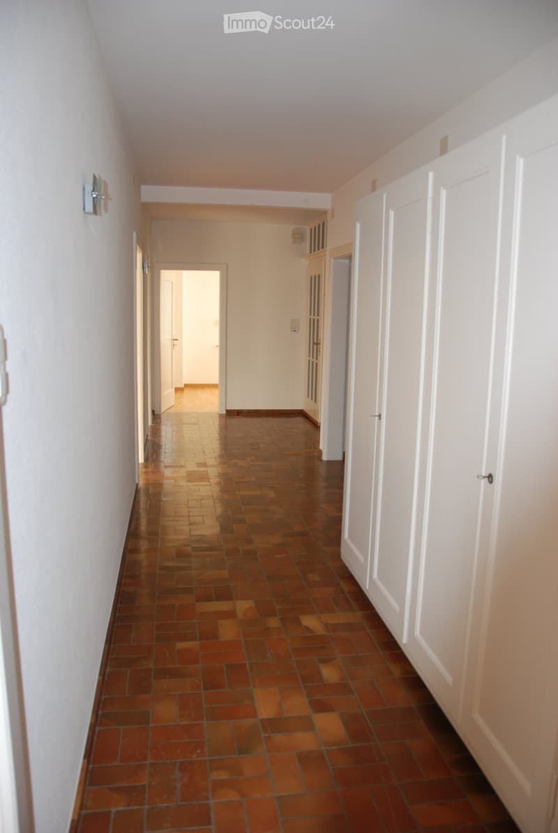 A long, narrow hallway with brown tiled flooring and white walls, featuring several closed doors and a visible light fixture on the wall.