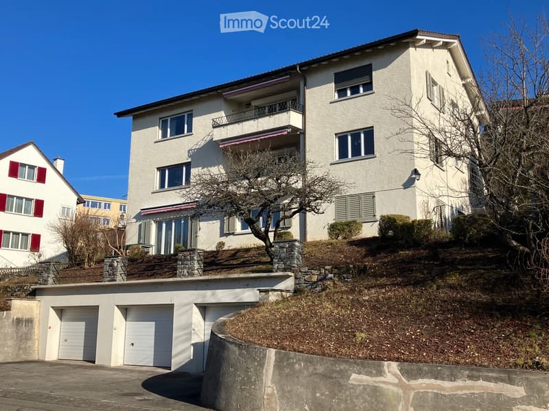 A multi-story white house with balconies, large windows, and a garage. The house is situated on a hillside with a stone retaining wall and a driveway leading to the garage.