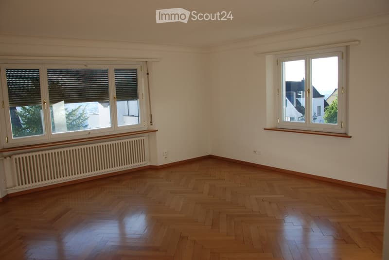 Empty room with wooden flooring, white walls, large windows with blinds, and radiators beneath the windows.