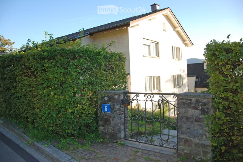 A two-story house with white walls and multiple windows, partially obscured by greenery and a stone gate with the number 6.
