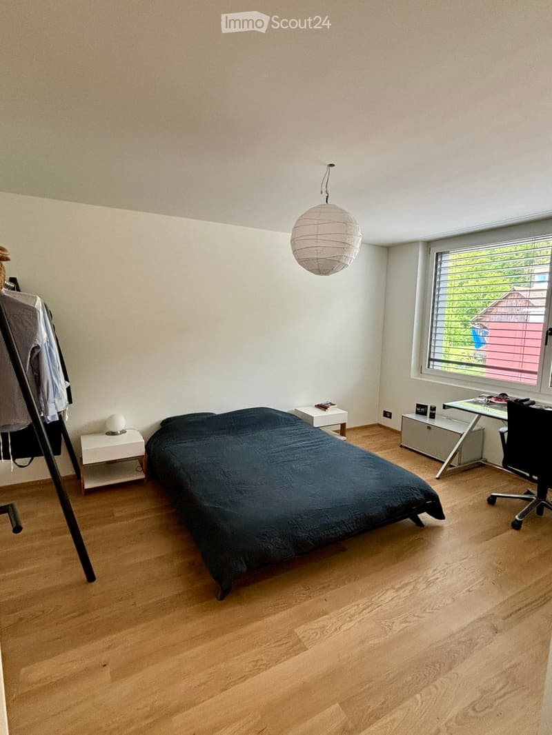 A modern bedroom with a wooden floor, a large bed with dark bedding, a nightstand, a desk with a chair, and a hanging paper lantern light fixture.