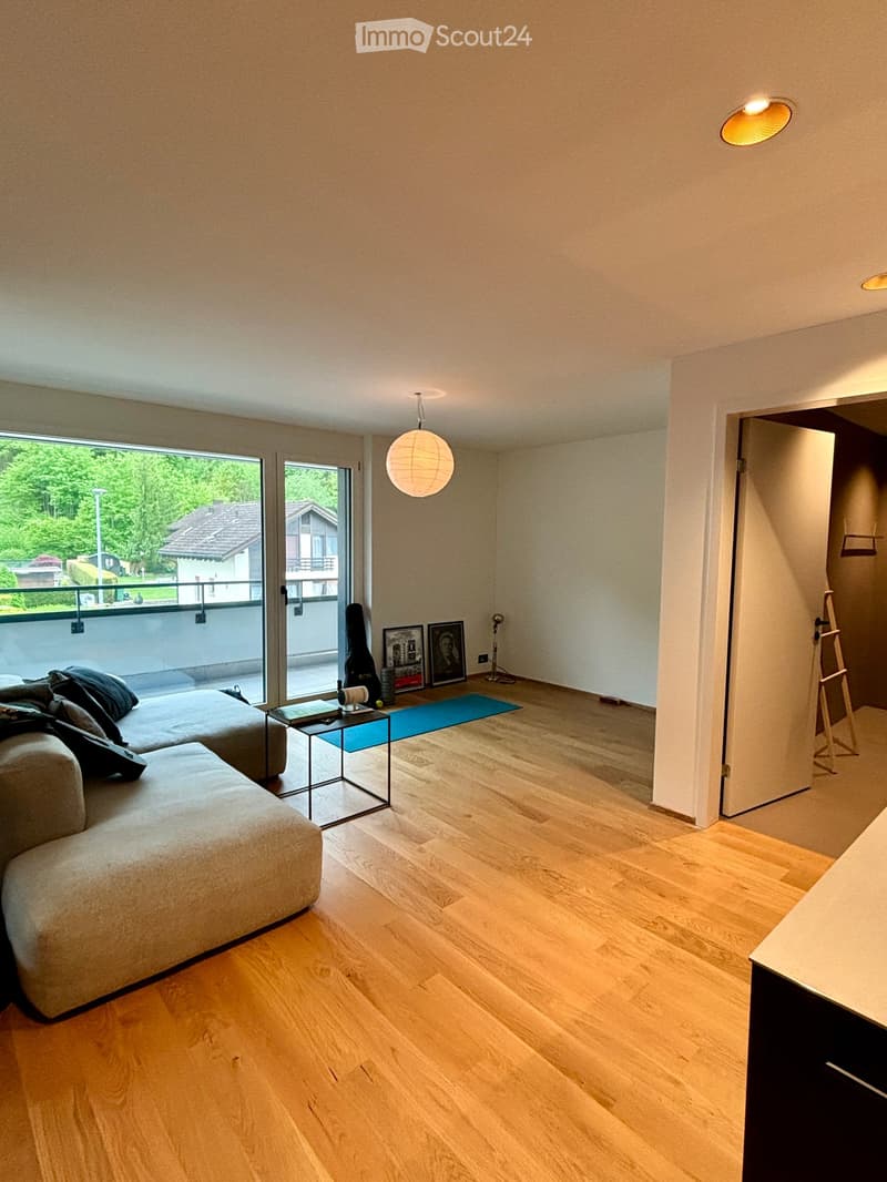 Modern living room with light wooden flooring, large windows, and a balcony. Features a grey sectional sofa, small glass table, framed pictures, and a guitar case.
