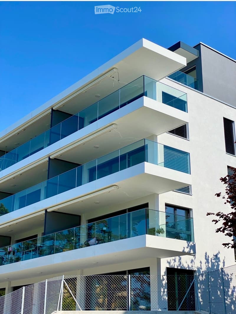 Modern multi-story building with white facade, large glass windows, and balconies with glass railings.