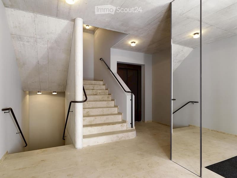 Modern hallway with a staircase leading upwards, featuring a sleek black metal railing, white walls, and recessed lighting.