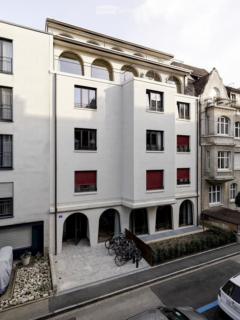 Modern multi-story building with white facade, arched windows, and balconies. Bicycles parked in front, small garden area with potted plants, and a covered car parked nearby.