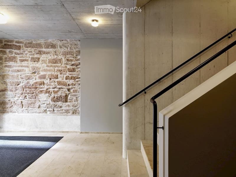 Modern hallway with a staircase featuring a black metal railing, beige tiled floor, and exposed brick wall on one side.